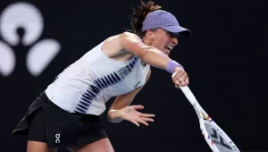 Tenisistka w białej koszulce z fioletowym daszkiem dynamicznie uderza piłkę rakietą podczas meczu tenisowego. Ekspresja twarzy zdradza pełne skupienie i determinację. Na tle rozmazane logo na tablicy reklamowej.