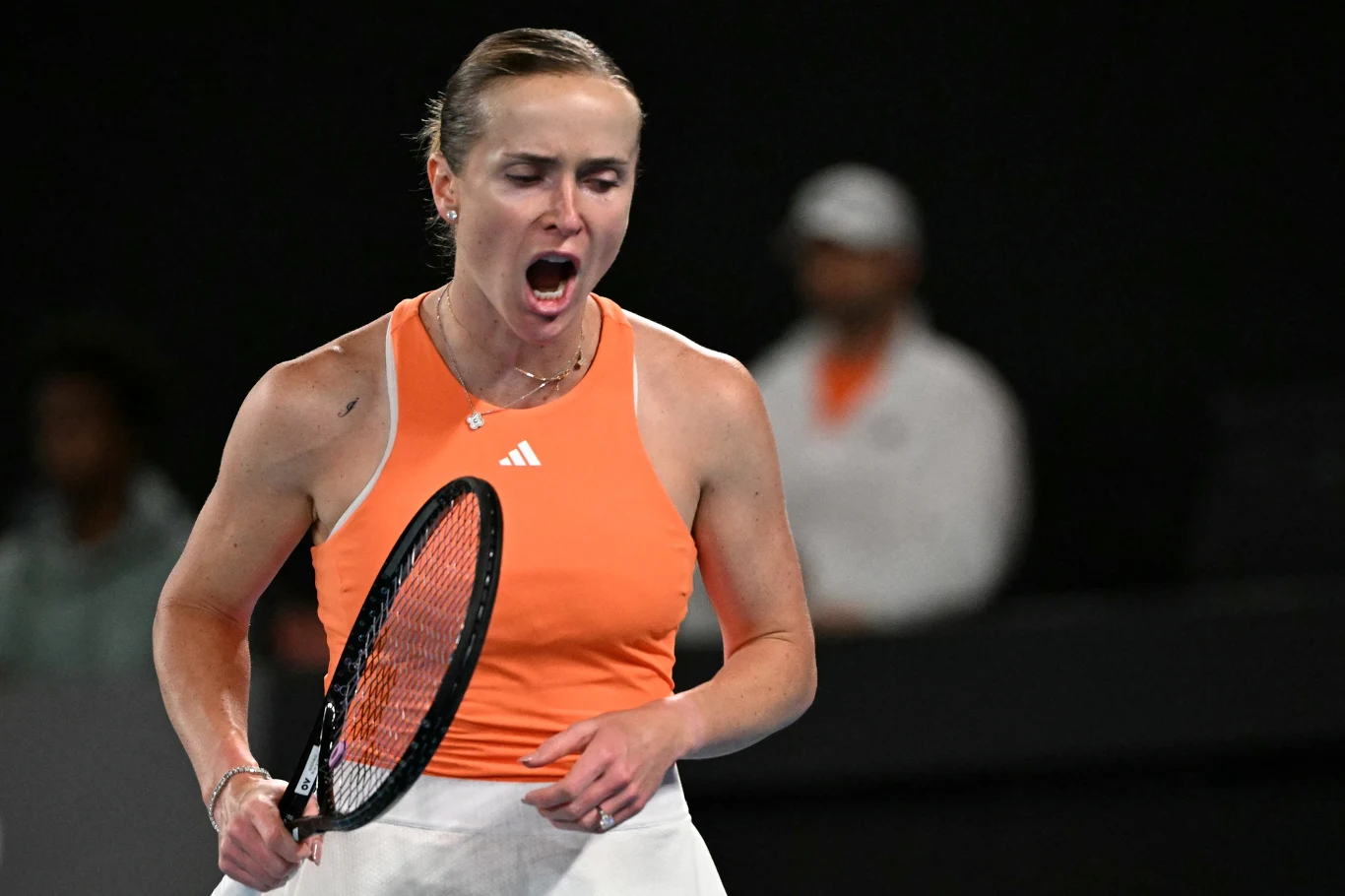 Zawodniczka tenisowa w pomarańczowym topie i białej spódnicy z rakietą w ręce wyraża silne emocje podczas meczu, w tle rozmyta postać w białym stroju.