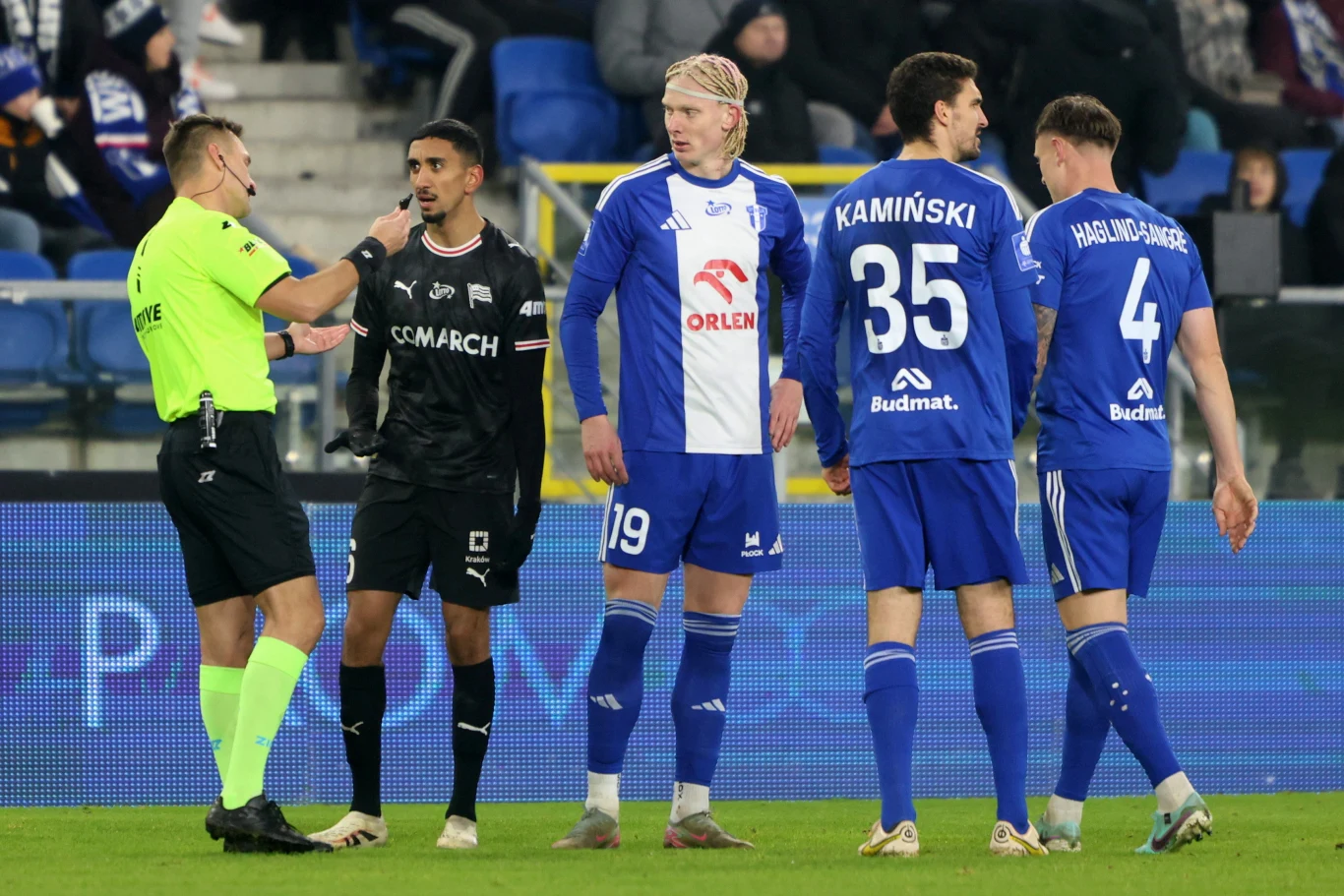 Sędzia piłkarski w jasnożółtym stroju rozmawia z zawodnikiem drużyny w czarnych strojach, obok stoją trzej piłkarze drużyny w niebiesko-białych koszulkach na murawie stadionu, w tle widać trybuny.