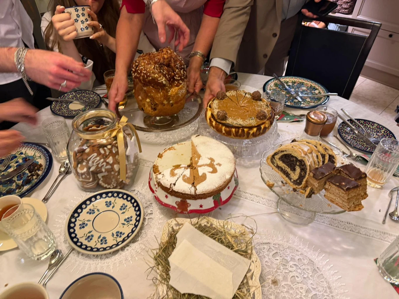 Zastawiony stół z tradycyjnymi ciastami, ciastkami i napojami, wokół którego siedzi kilka osób, sięgających po słodycze. Na talerzach widoczne są wypieki, między innymi makowiec, sernik, piernik oraz słoik z dekoracyjnym motywem.