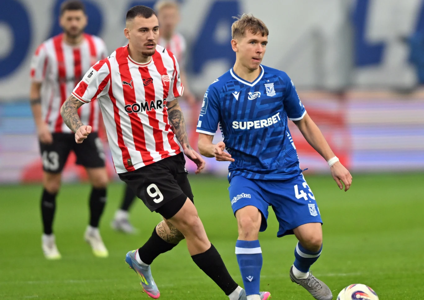 Dwóch piłkarzy walczących o piłkę podczas meczu, jeden zawodnik w biało-czerwonym pasiastym stroju drużyny Cracovii oraz drugi w niebieskim stroju Lecha Poznań na zielonym boisku.