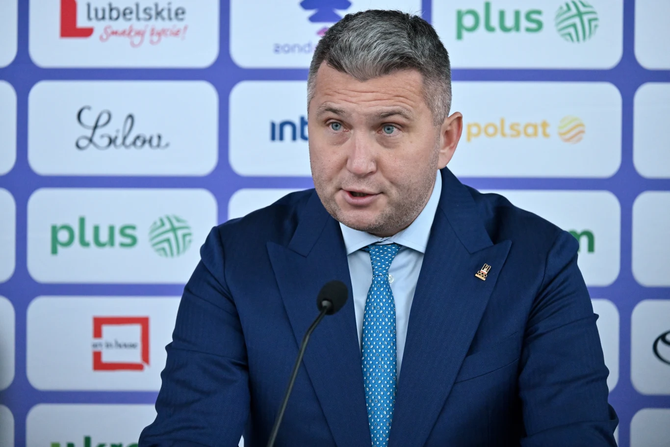 Mężczyzna w garniturze przemawia do mikrofonu podczas konferencji prasowej, w tle widoczne logotypy sponsorów i partnerów wydarzenia.