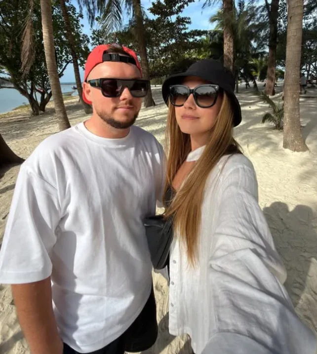 Para młodych ludzi w okularach przeciwsłonecznych pozuje razem na piaszczystej plaży w otoczeniu palm. Kobieta ubrana w jasną koszulę i czarny kapelusz, mężczyzna w białej koszulce i czerwonej czapce z daszkiem. Słoneczna pogoda, w tle widoczne drzewa ...