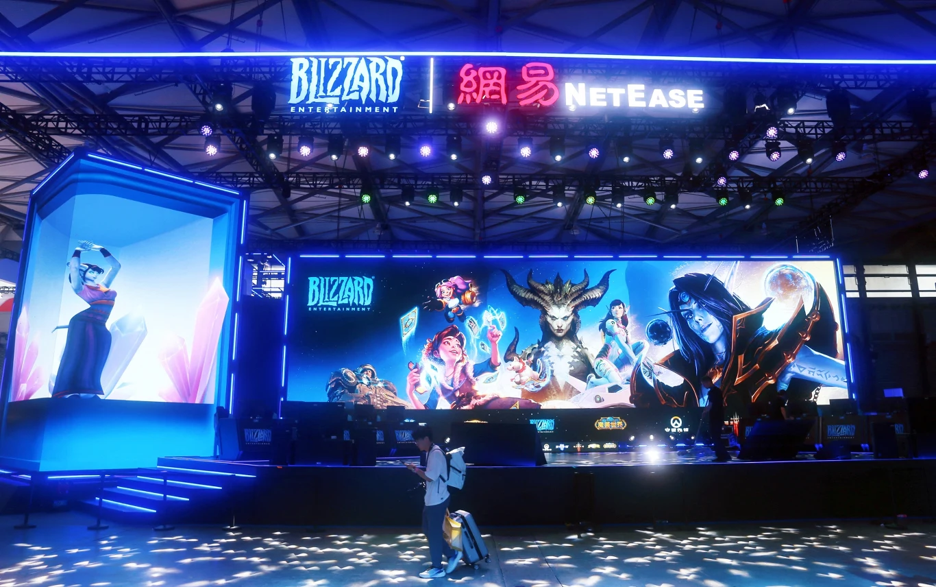 Nadciąga Blizzard Showcase Duża scena z kolorowymi światłami i ogromnym ekranem, na którym widoczne są postacie z gier komputerowych firmy Blizzard Entertainment. Logo Blizzard i NetEase na górze stoiska podkreślają współpracę obu firm. Przed sceną pojedyncza osoba robi zdjęcie,...