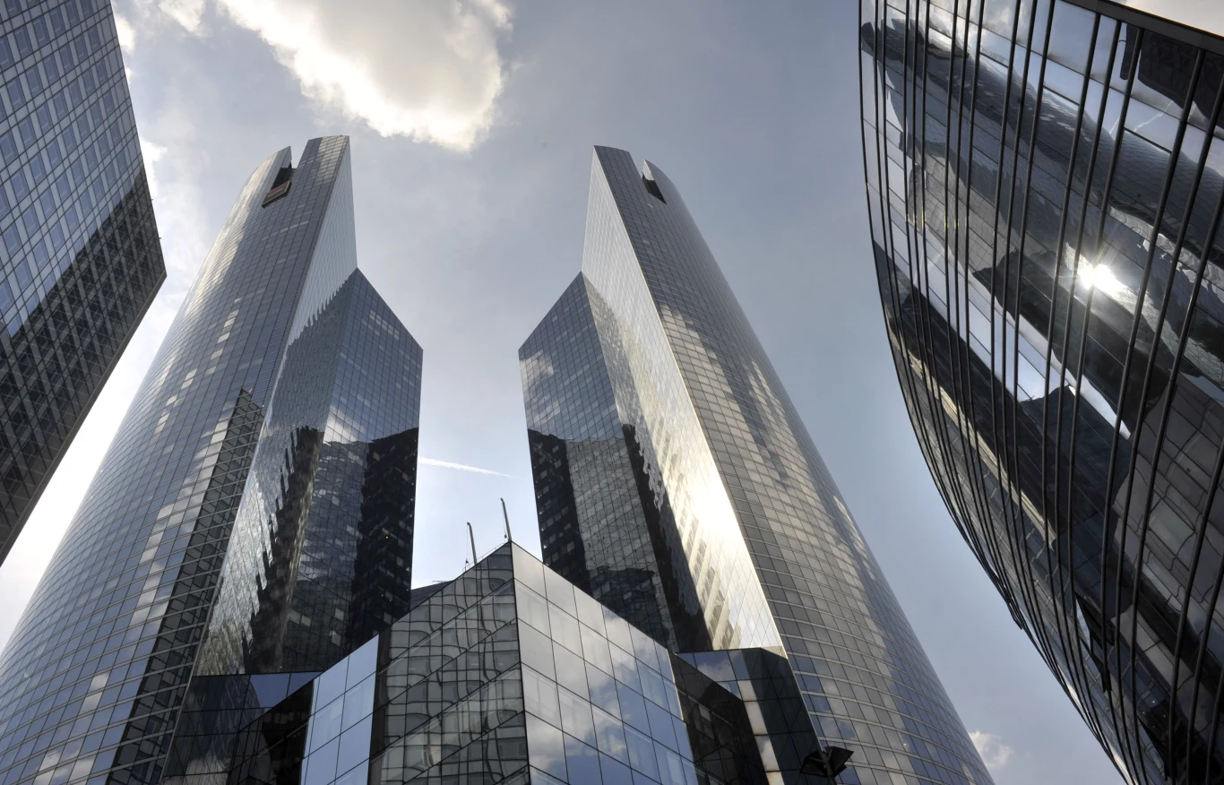 La Defense w Paryżu. Dzielnica biznesowa, która świeci pustkami Wysokie, nowoczesne wieżowce o przeszklonych elewacjach, odbijające światło słoneczne. Budynki stoją blisko siebie, dominując nad okolicą. Na niebie widoczna chmura oraz smuga po samolocie, całość utrzymana w chłodnych tonacjach.