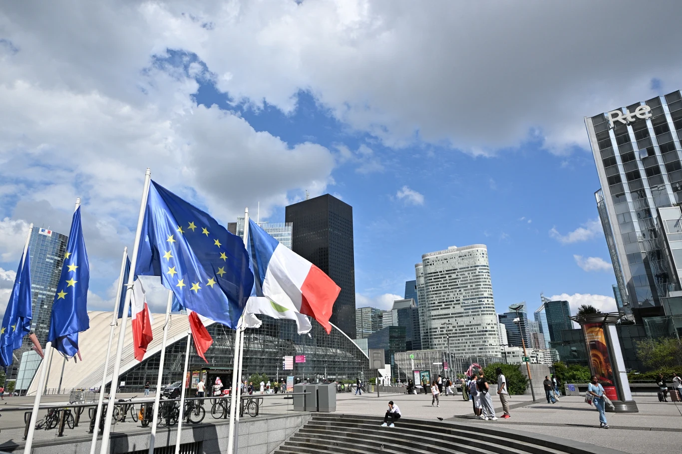 Dzielnica La Defense w Paryżu Flagi Unii Europejskiej i Francji rozwiewają się na wietrze przed nowoczesnymi wieżowcami dzielnicy biznesowej La Défense w Paryżu, pośród przechodniów oraz rowerzystów na tle nieba z licznymi chmurami.