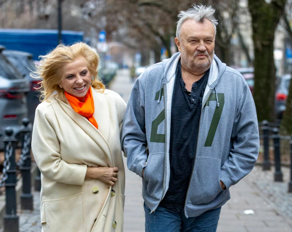 Joanna i Krzysztof Cugowscy są razem od połowy lat 90. Dwie uśmiechnięte osoby spacerujące chodnikiem w miejskim otoczeniu. Kobieta w jasnym płaszczu i pomarańczowym szaliku trzyma ręce przed sobą, mężczyzna ubrany w szarą bluzę i ciemny t-shirt idzie obok niej. Drzewa bez liści oraz zamazane samochody i o...