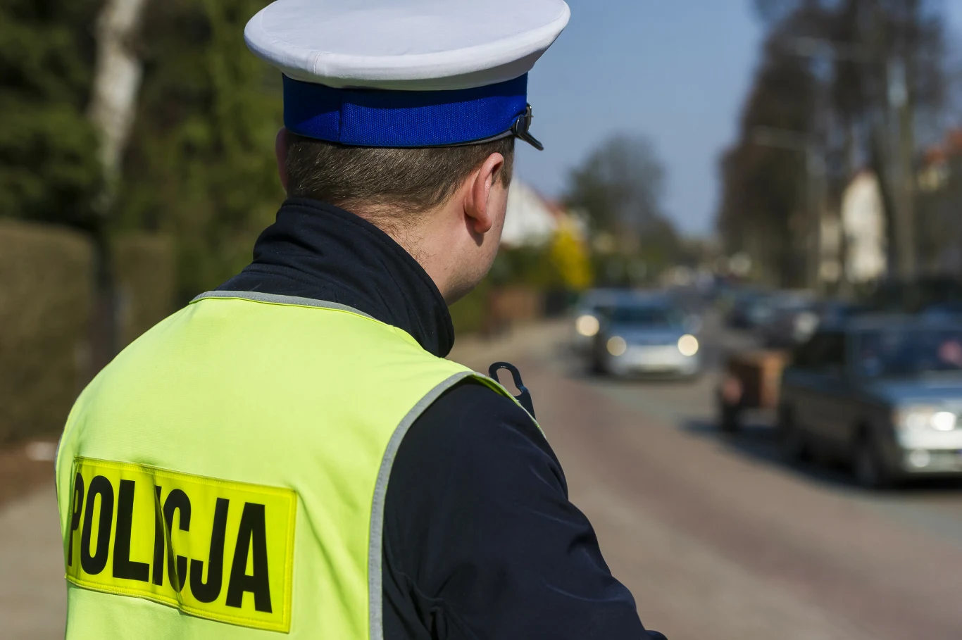 Policjant coraz częściej zagląda podczas kontroli do wnętrza pojazdu albo bagażnika. Funkcjonariusz policji w żółtej kamizelce z napisem 'Policja' stoi przy drodze, obserwując przejeżdżające samochody w słoneczny dzień, w tle widoczne są drzewa i zabudowania mieszkalne.