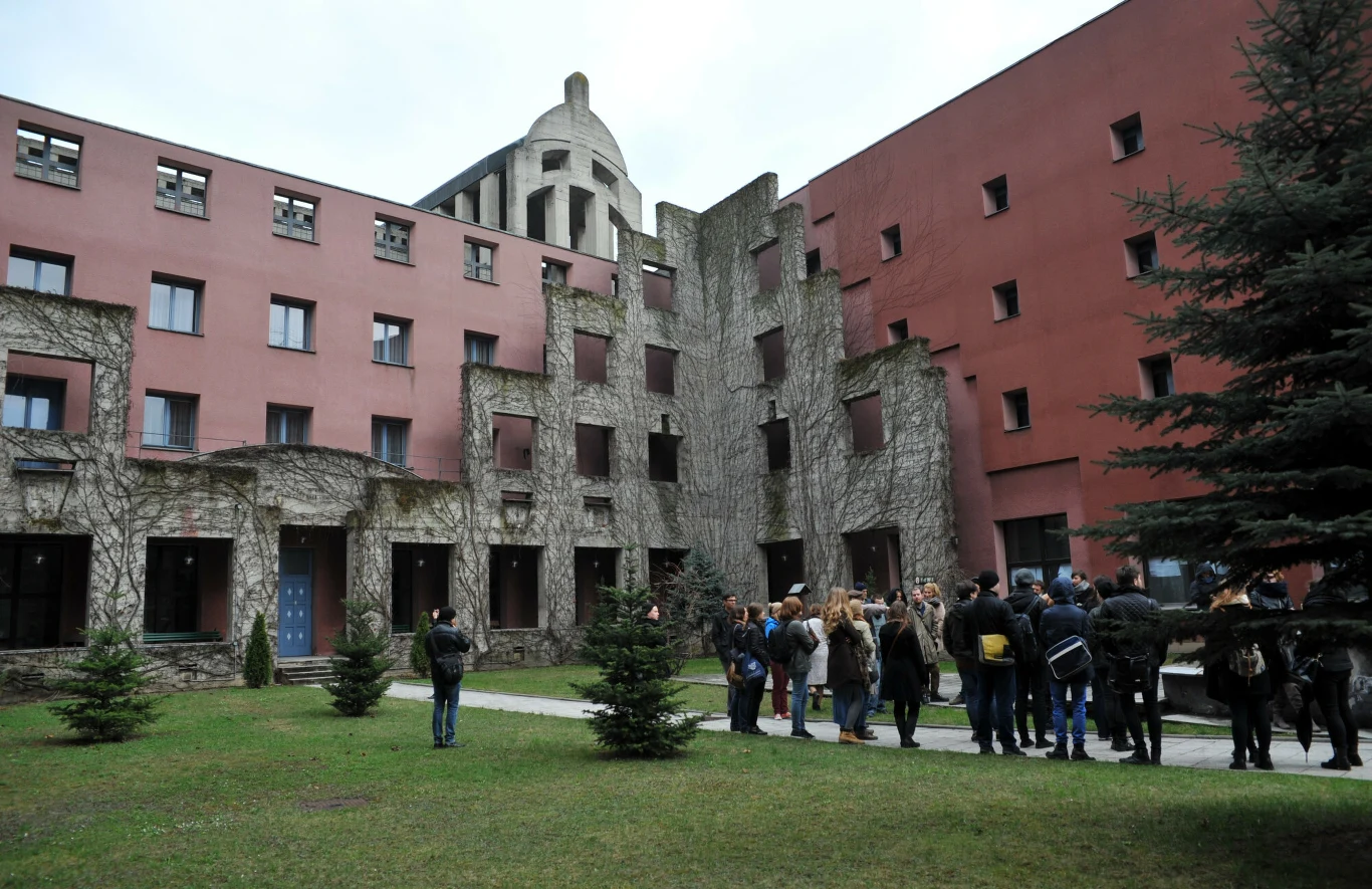 Grupa ludzi zgromadzona przed charakterystycznym budynkiem z czerwonymi i szarymi ścianami, ozdobionym pnączami, na tle którego widać oryginalnie wyglądającą kopułę. Wokół znajduje się trawnik oraz kilka drzewek.