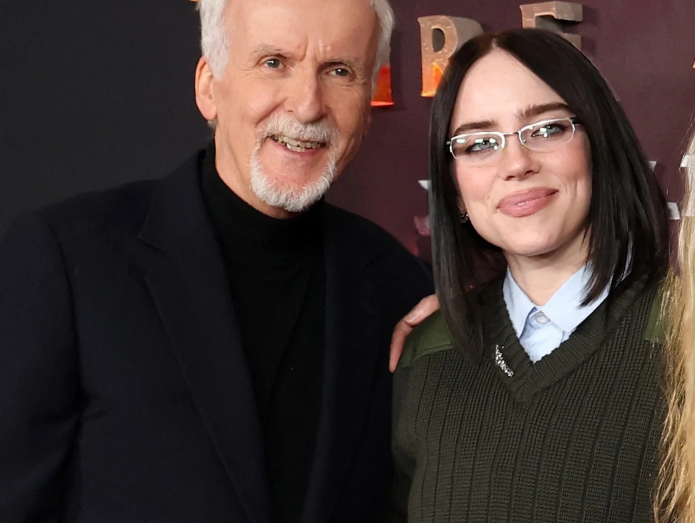 James Cameron i Billie Eilish stworzyli film koncertowy Dwie osoby stojące obok siebie, mężczyzna o siwych włosach w czarnym garniturze i kobieta z ciemnymi włosami w okularach, ubrana w zielony sweter i niebieską koszulę, uśmiechnięci, na tle nieczytelnego napisu.