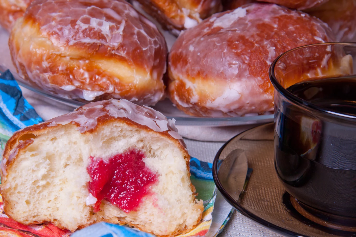 W tłusty czwartek trzeba zjeść przynajmniej jednego pączka Pączki z lukrem wypełnione czerwonym dżemem, na pierwszym planie przekrojony pączek odsłaniający nadzienie, obok filiżanka czarnej kawy.