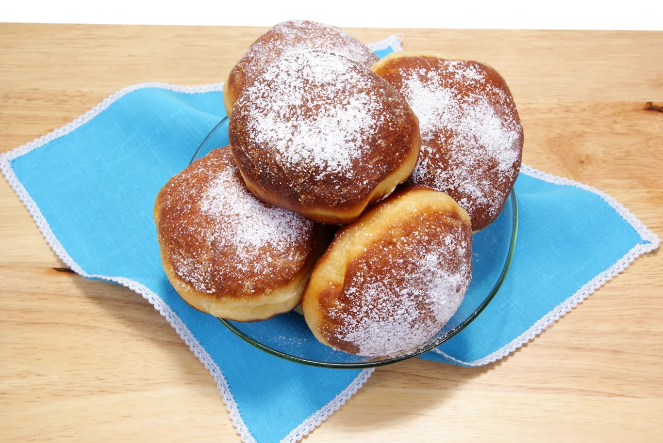 Stos pączków oprószonych cukrem pudrem ułożony na szklanym talerzu, umieszczonym na jasnoniebieskiej serwetce, na drewnianym blacie.