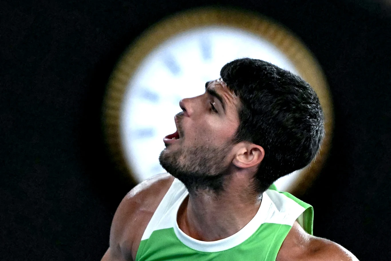 Sportowiec w zielono-białym stroju, z widocznym napięciem mięśni i wyrazem intensywnych emocji na twarzy, na tle rozmytego zegara.