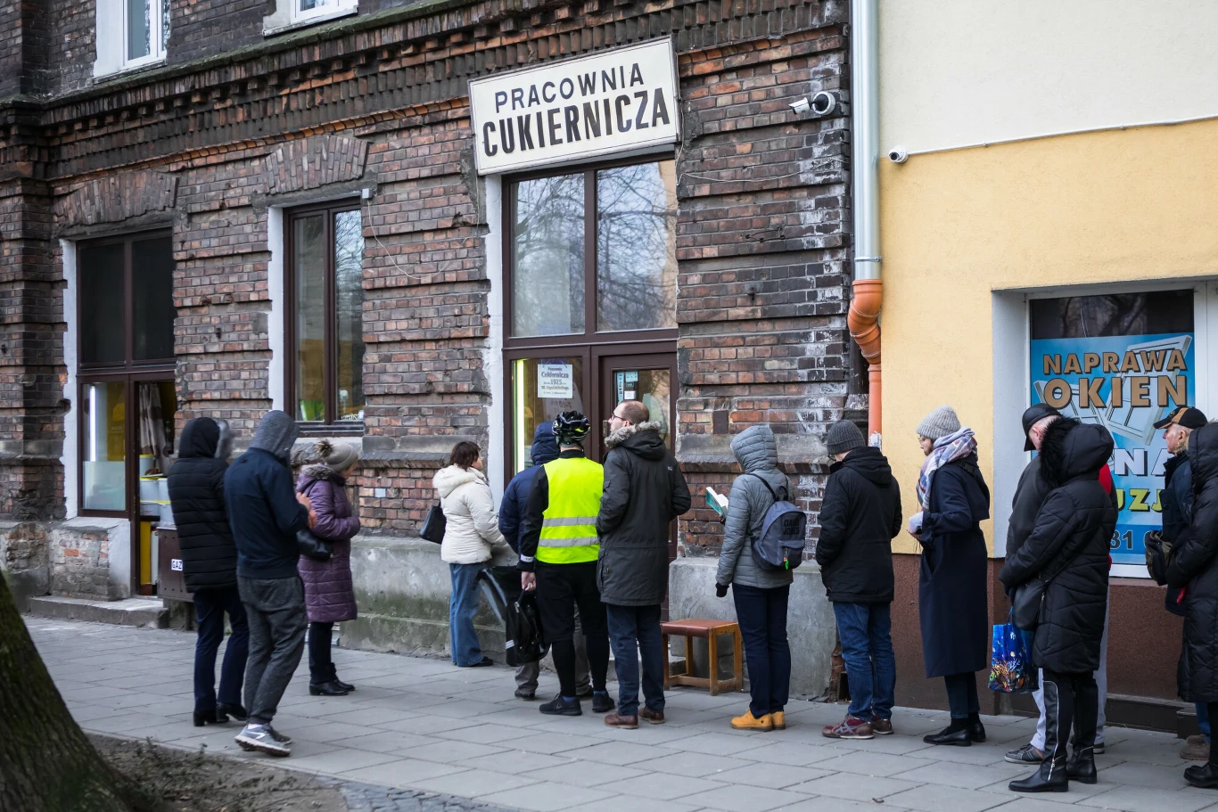 Grupa osób ubranych w zimowe ubrania stoi w kolejce pod cukiernią w starym budynku z cegły, przy wejściu znajduje się osoba w żółtej kamizelce odblaskowej.