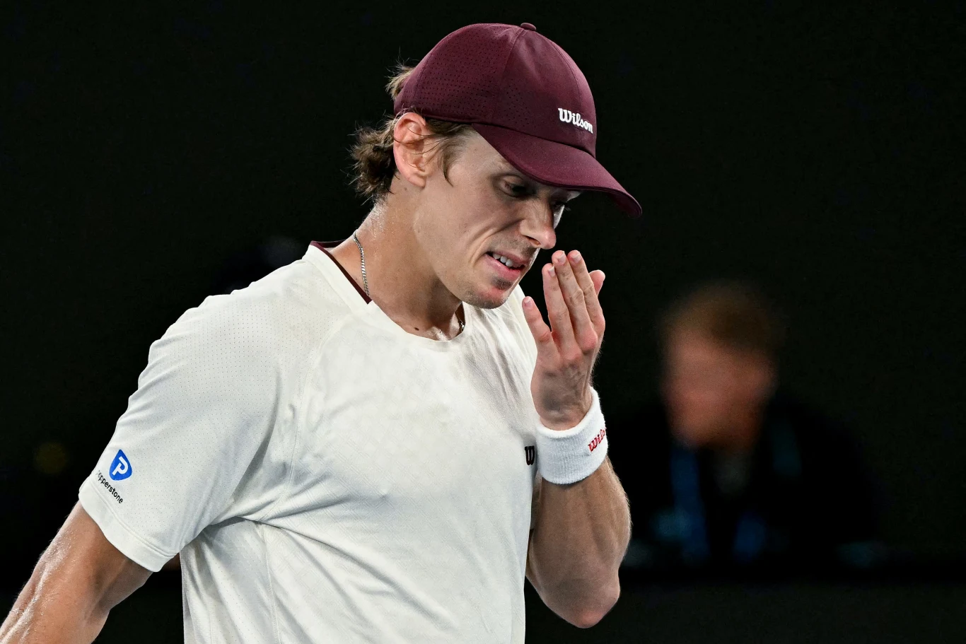 Młody mężczyzna w sportowej koszulce i bordowej czapce z daszkiem ociera dłoń o twarz, stojąc na korcie tenisowym podczas meczu. Jego wyraz twarzy wskazuje na skupienie lub zmęczenie.