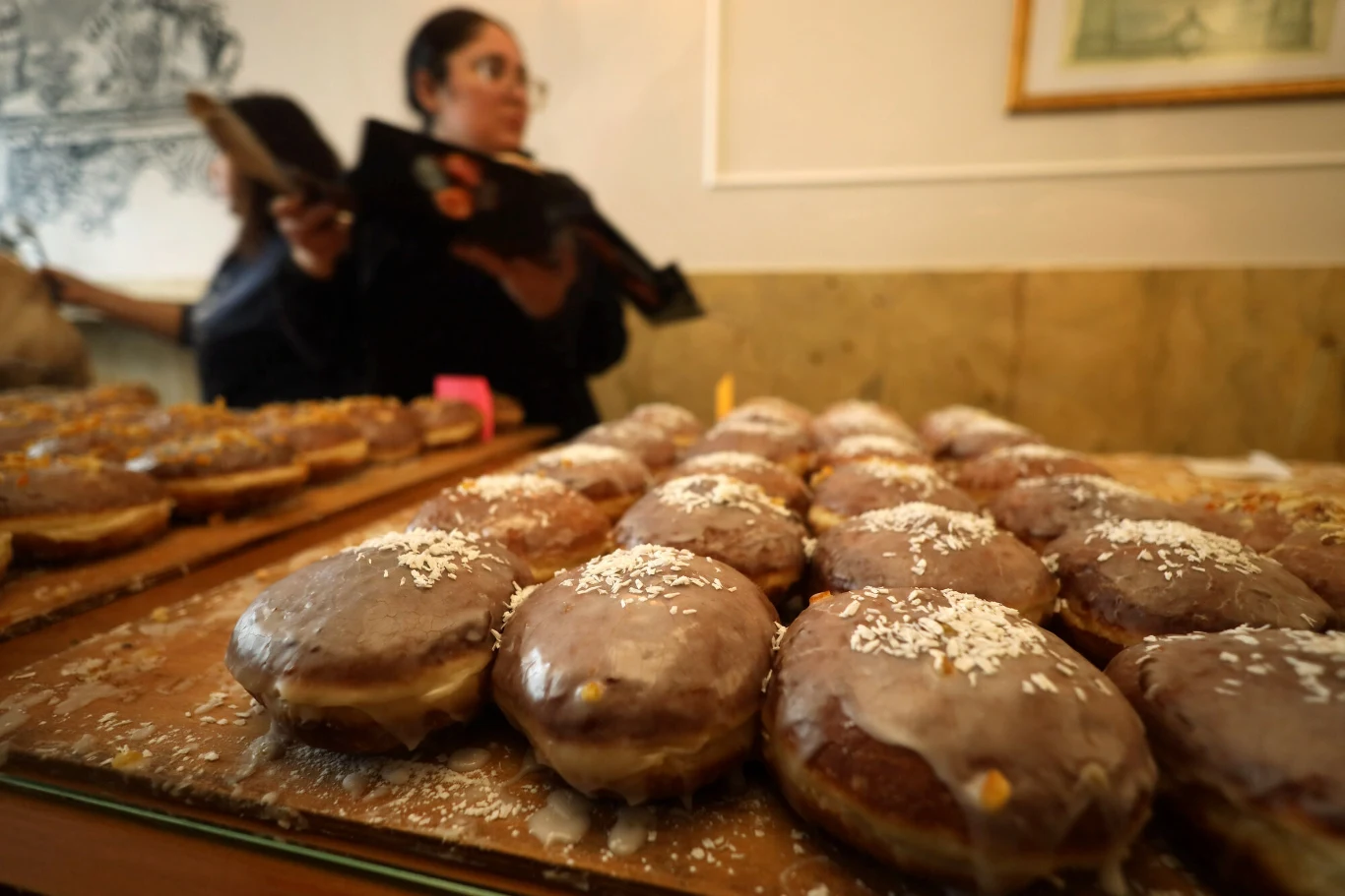 Rząd pączków z lukrem i posypką na blacie w cukierni, w tle kobieta trzymająca tacę.