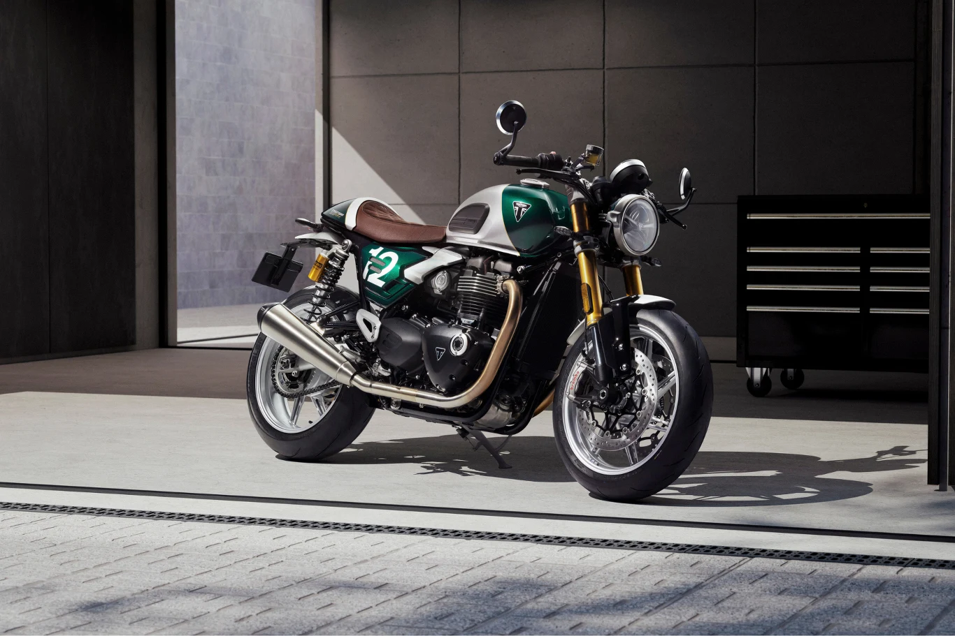 Limitowany Triumph Speed Twin 1200 Cafe Racer Edition - powstanie zaledwie 800 egzemplarzy. Motocykl w stylu retro z numerem 72 na baku, lakierowany na zielono z brązową skórzaną kanapą, stoi na tle nowoczesnego warsztatu z czarną szafką narzędziową.