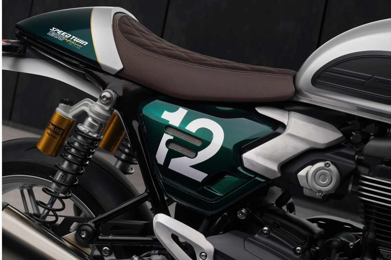 Speed Twin 1200 Cafe Racer Edition to hołd dla cafe racerów z lat 60. Boczny widok zielonego motocykla z białym numerem 12 na metalowej części, brązowe siedzenie oraz widoczne elementy tylnego zawieszenia i amortyzatora.