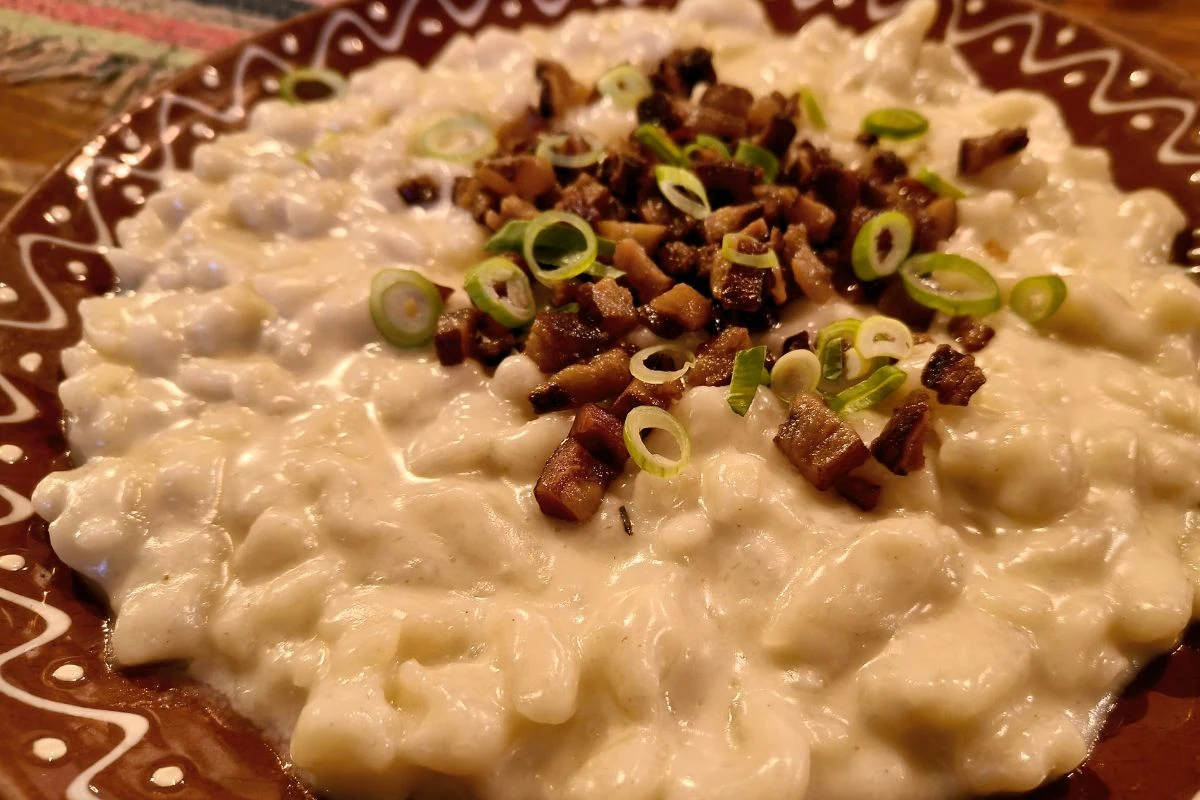 Tradycyjna potrawa składająca się z klusek w kremowym sosie, udekorowana podsmażonym boczkiem i zieloną cebulką na ozdobnym talerzu.