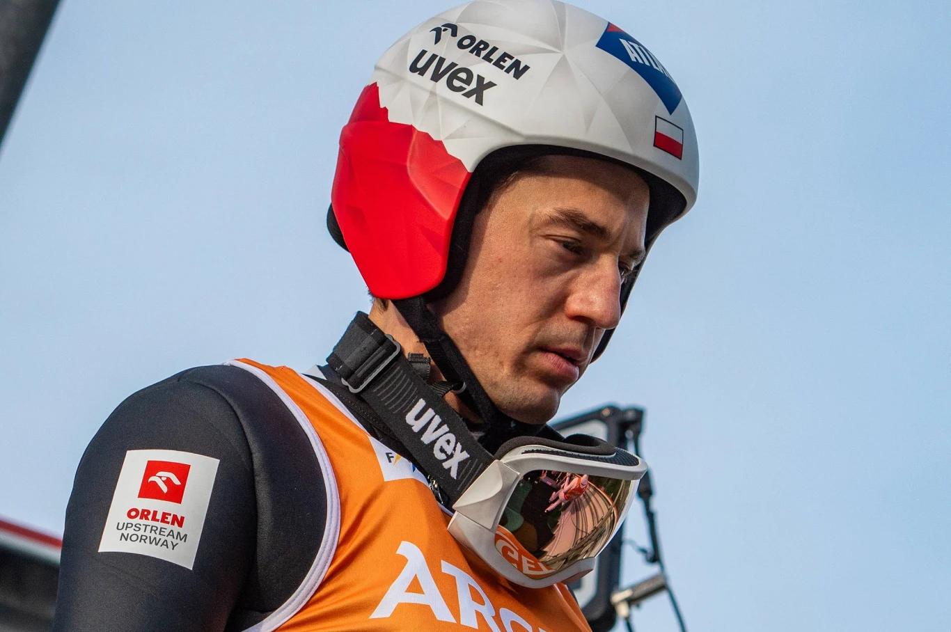 Kamil Stoch, chorąży reprezentacji Polski na zimowe igrzyska olimpijskie Mediolan-Cortina 2026 Sportowiec w kombinezonie narciarskim, z kaskiem i goglami na szyi, ubrany w pomarańczową kamizelkę z logotypami sponsorów, skupiony przed startem