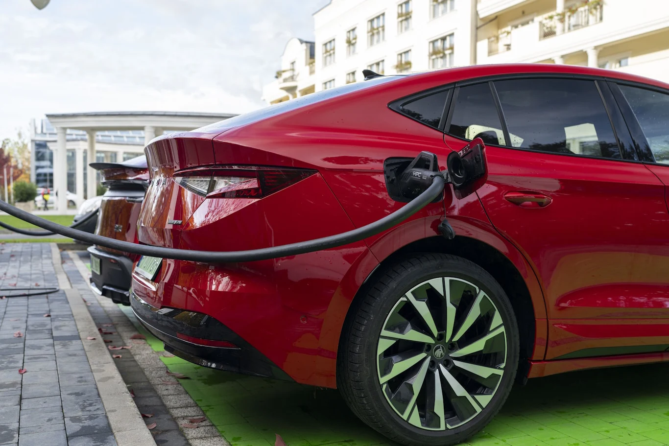 Czerwony samochód elektryczny podczas ładowania na zielonym stanowisku przy miejskim budynku, widoczne złącze ładowarki podpięte do portu ładowania samochodu.