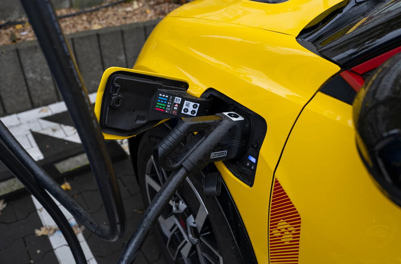 Ile kosztuje ładowanie auta elektrycznego w Polsce? Żółty samochód elektryczny stojący przy stacji ładowania, przewód ładowarki włożony do gniazda pojazdu, widoczne szczegóły techniczne panelu ładowania oraz fragment koła.