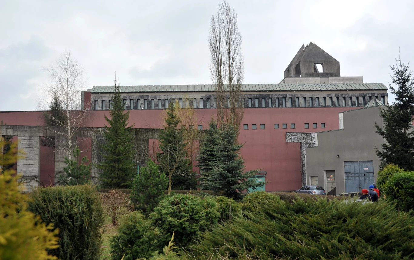 Nowoczesny, masywny budynek z niestandardowym geometrycznym dachem, otoczony zielonymi krzewami i drzewami iglastymi. W tle pochmurne niebo, a przy ścianie budynku widoczne są drzwi i fragmenty okien.