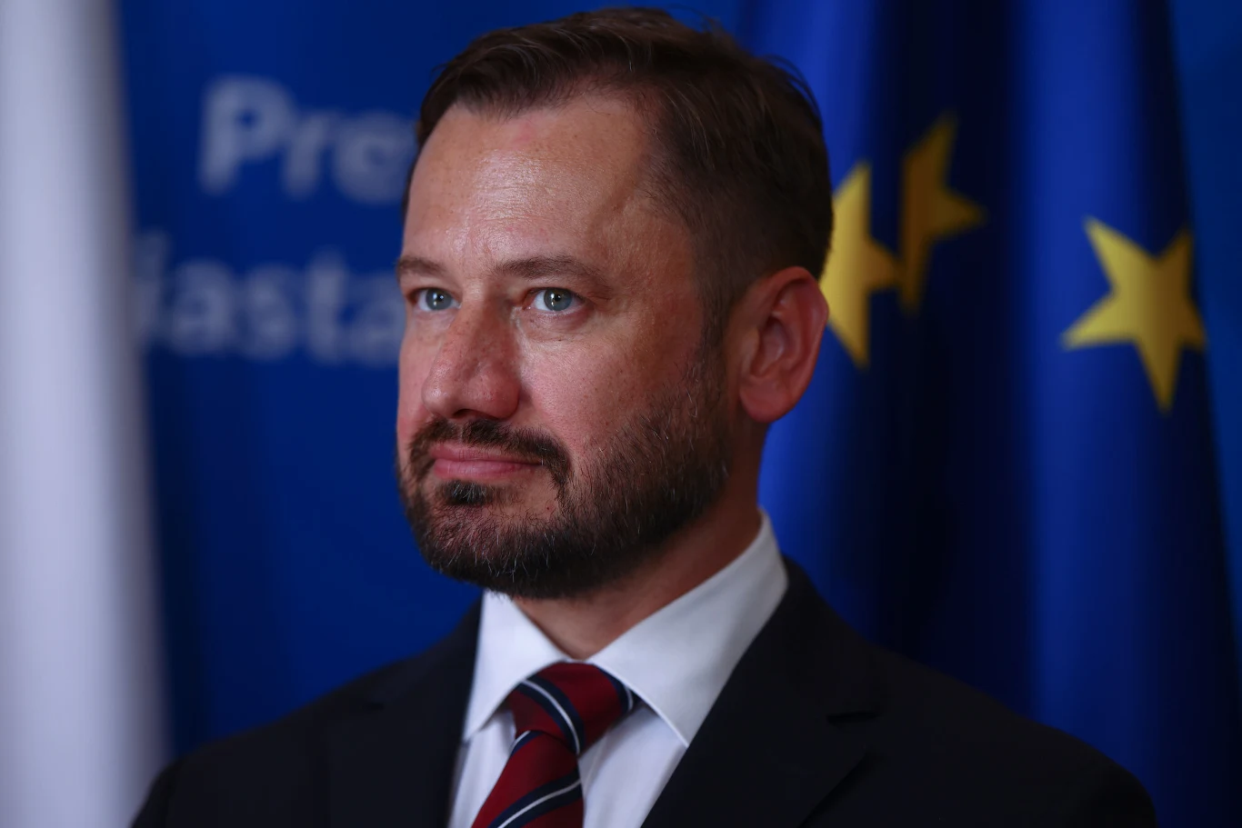 Mężczyzna w eleganckim garniturze i krawacie, z krótką brodą, stoi na tle niebieskich flag ze złotymi gwiazdami oraz białej flagi, jego wyraz twarzy jest poważny i skupiony.
