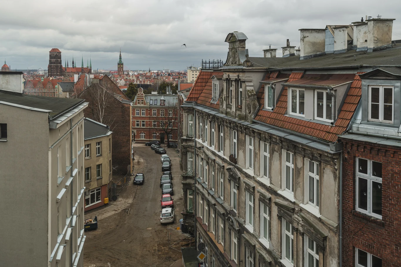 Kamienice z cegły i tynku ustawione wzdłuż wąskiej uliczki, szereg zaparkowanych samochodów, w tle panorama miasta z zabytkowymi wieżami kościołów pod zachmurzonym niebem.