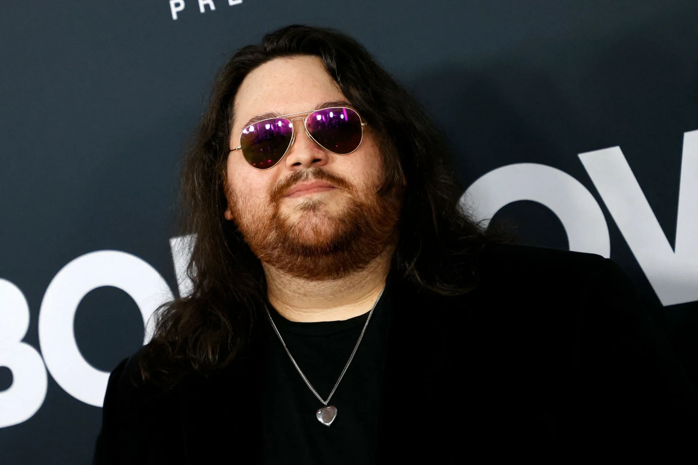 Wolfgang Van Halen powraca do Polski z projektem Mammoth Mężczyzna o długich ciemnych włosach i brodzie, w okularach przeciwsłonecznych z różowymi szkłami, ubrany w ciemną marynarkę i czarną koszulkę z naszyjnikiem na szyi, stoi na tle ciemnej ścianki z dużymi białymi literami.