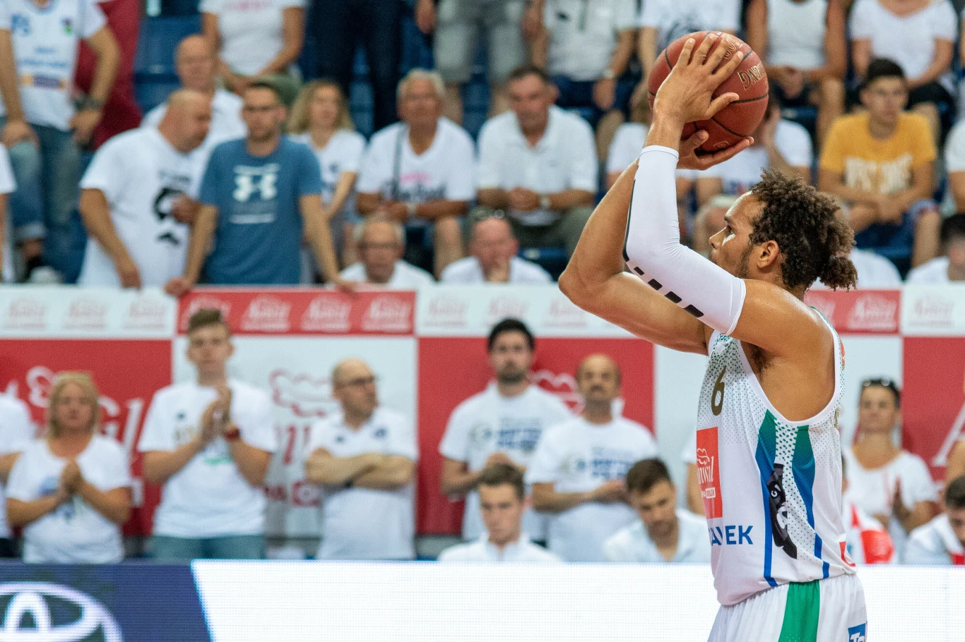 Koszykarz przygotowujący się do rzutu wolnego na boisku podczas meczu, w tle widoczna publiczność ubrana w białe koszulki kibiców.