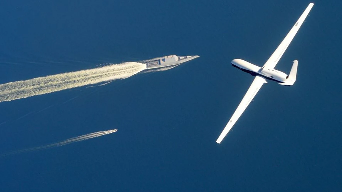 Szpiegowskie drony USA i Iranu minęły się w powietrzu Duży bezzałogowy samolot wojskowy przelatuje nad morzem w pobliżu nowoczesnego okrętu wojennego. W oddali widoczna jest motorówka oraz smuga wody pozostawiona na powierzchni przez okręt.