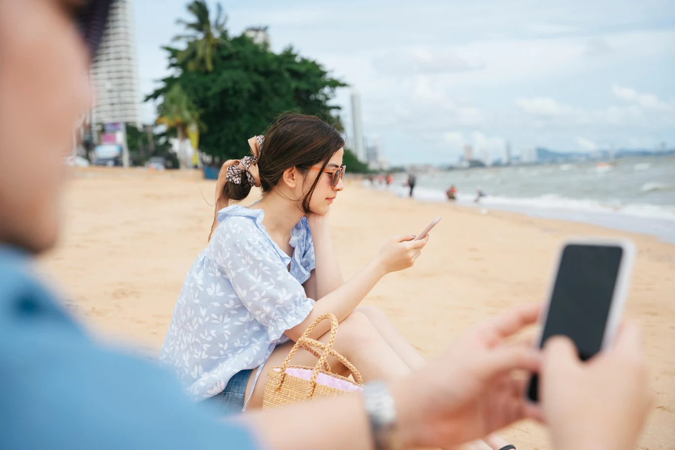 Młoda kobieta siedzi na plaży z telefonem w dłoni, obok niej leży torba. W tle rozległe morze, piasek oraz palmy. W lewej części zdjęcia widoczna jest niewyraźna postać trzymająca telefon.