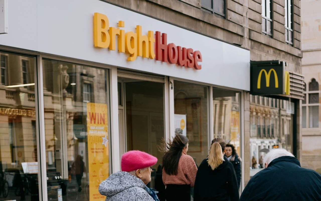 Grupa ludzi idących wzdłuż ulicy przed witryną sklepu BrightHouse oraz pobliską restauracją McDonald's, widoczne są szyldy obu lokali.