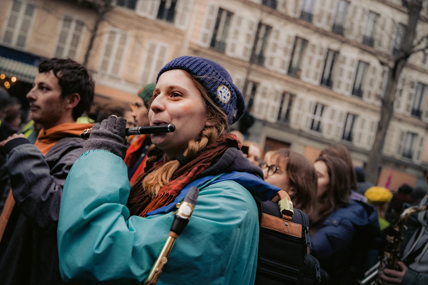 Francuski gigant tonie. Nadchodzi fala strajków Młoda kobieta w niebieskiej kurtce i czapce gra na flecie w tłumie ludzi na ulicy, wokół niej widoczne są skupione twarze innych uczestników wydarzenia oraz kamienice w tle.