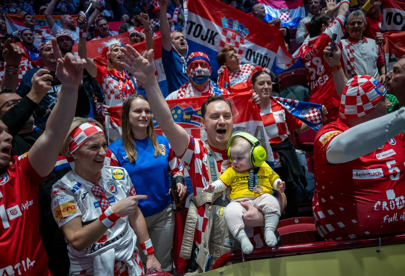 Tłum chorwackich kibiców celebrujących z flagami i szalikami, w centrum mężczyzna trzymający małe dziecko w żółtych słuchawkach ochronnych, wszyscy ubrani w barwy narodowe Chorwacji.