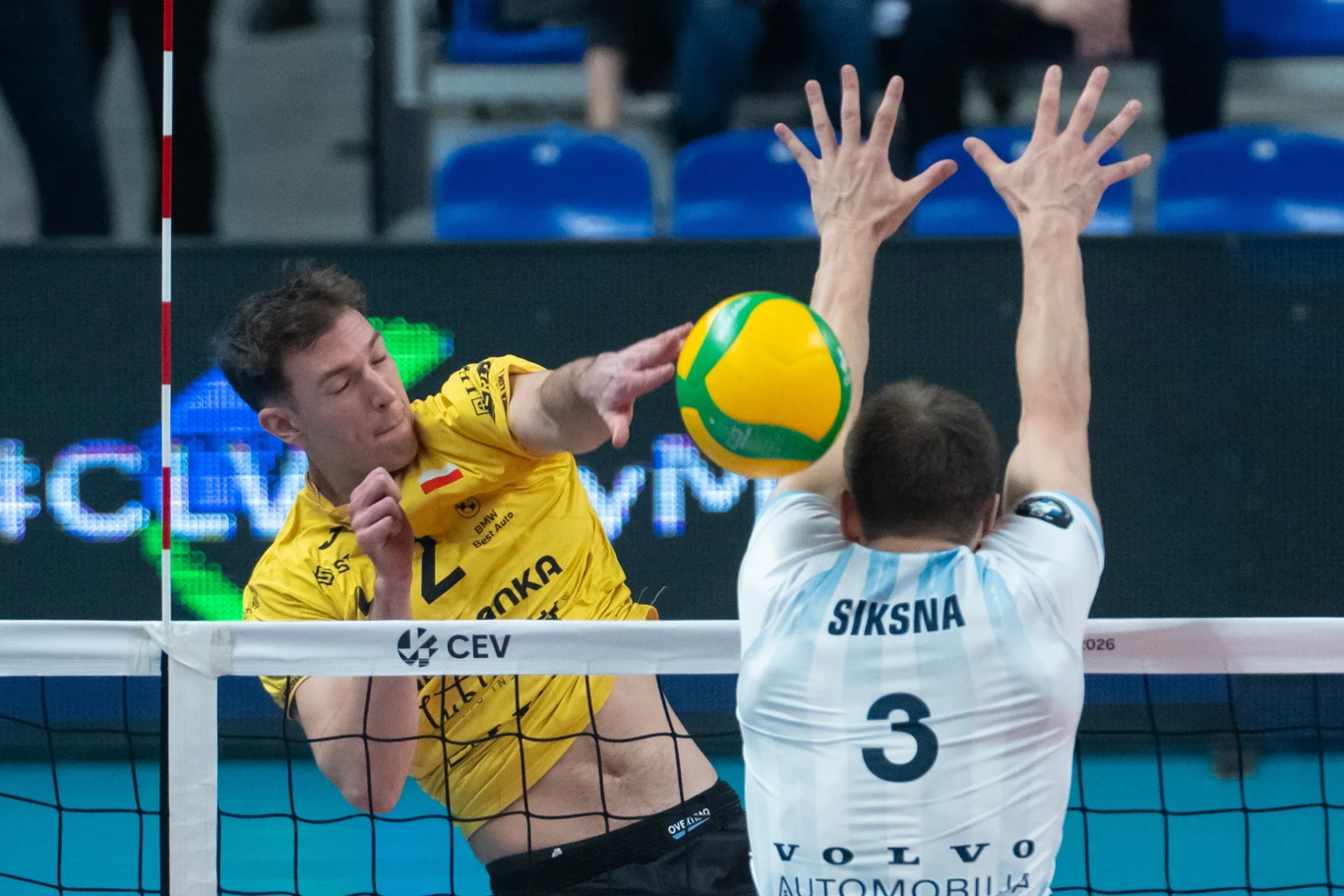 Zawodnik w żółtej koszulce atakuje piłkę do siatkówki nad siatką, naprzeciwko zawodnik w białej koszulce z numerem 3 unosi ręce w próbie bloku. W tle widoczne niebieskie krzesełka hali sportowej.