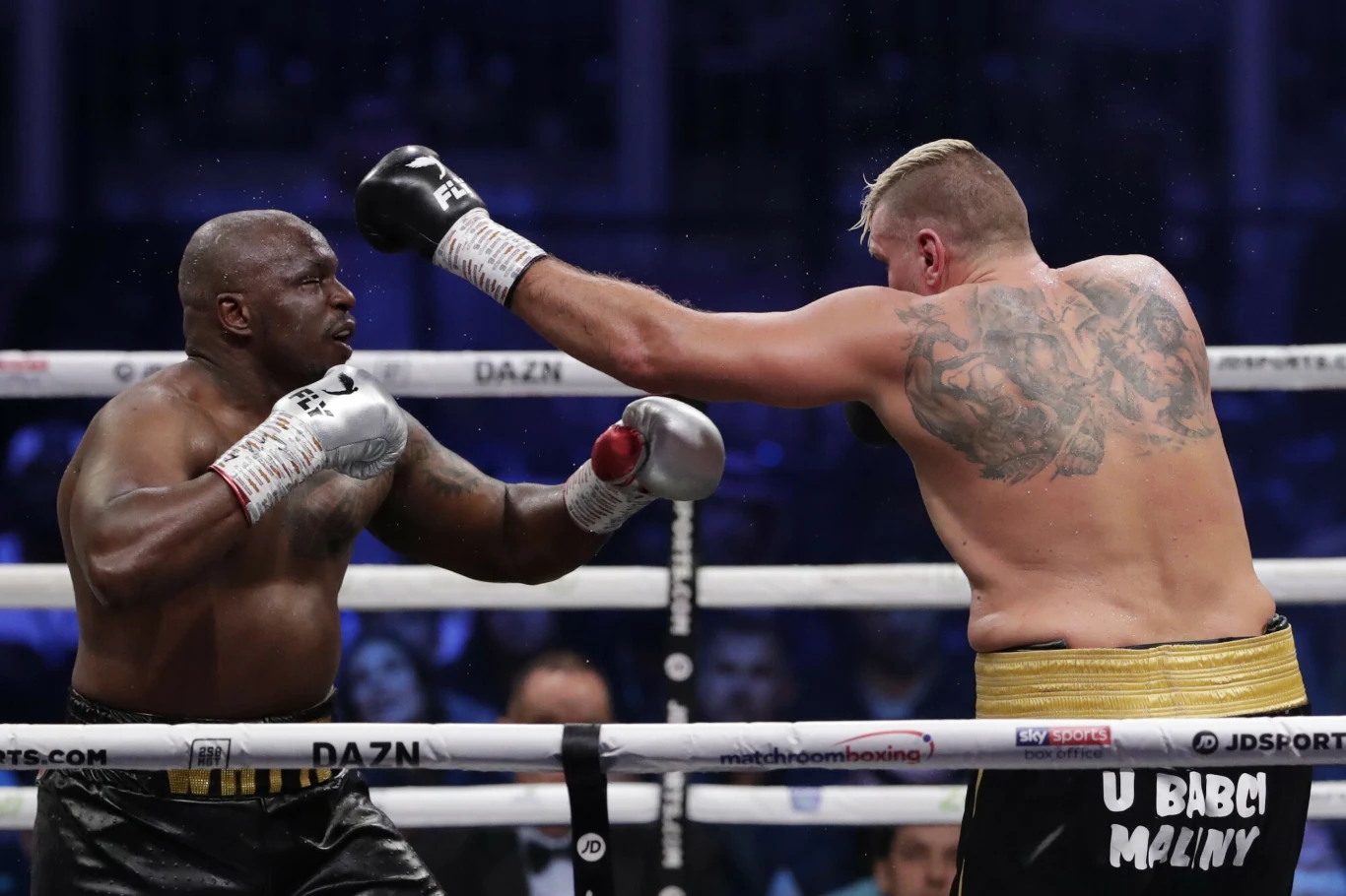 Dwóch bokserów zmierza się na ringu, jeden z nich zadaje dynamiczny cios, a drugi przygotowuje się do obrony; wyraźnie widoczne umięśnione sylwetki, tatuaże oraz rękawice bokserskie, w tle publiczność i barierki ringu.