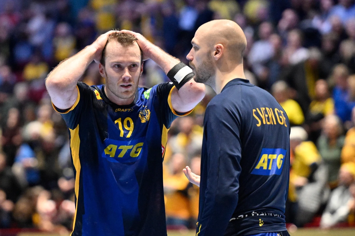 Dwóch zawodników w żółto-niebieskich strojach reprezentacji Szwecji, jeden z numerem 19, drugi z napisem 'Sverige' na plecach, rozmawiają ze sobą. Pierwszy z nich trzyma ręce na głowie i wydaje się zaniepokojony lub rozczarowany. W tle rozmyta publiczn...