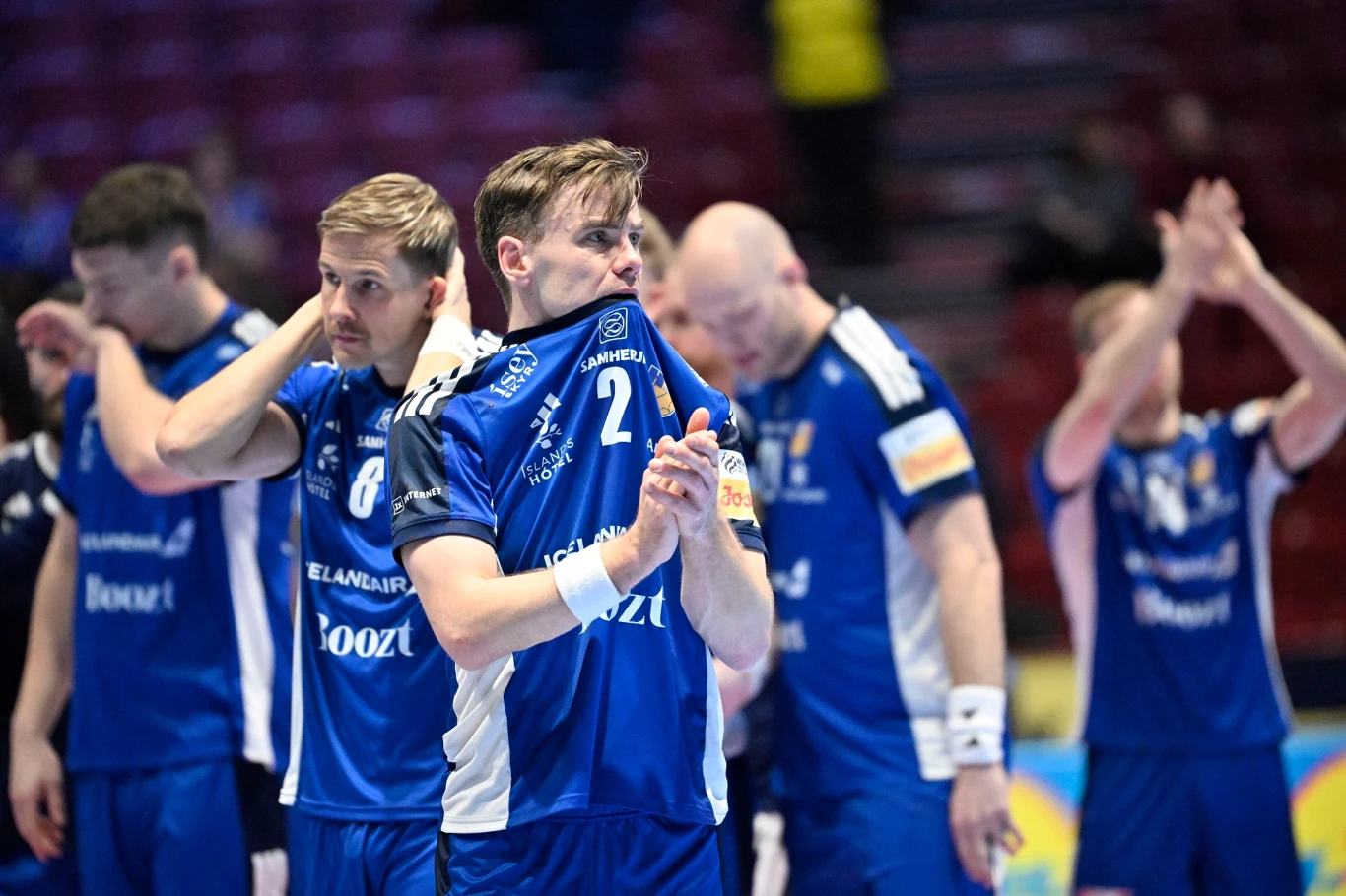 Grupa sportowców w niebieskich strojach stoi na boisku, okazując widoczne emocje i rozczarowanie po zakończonym meczu. Niektórzy mają głowy pochylone, jeden z zawodników zakrywa twarz koszulką, a inni klaszczą lub dotykają głowy.