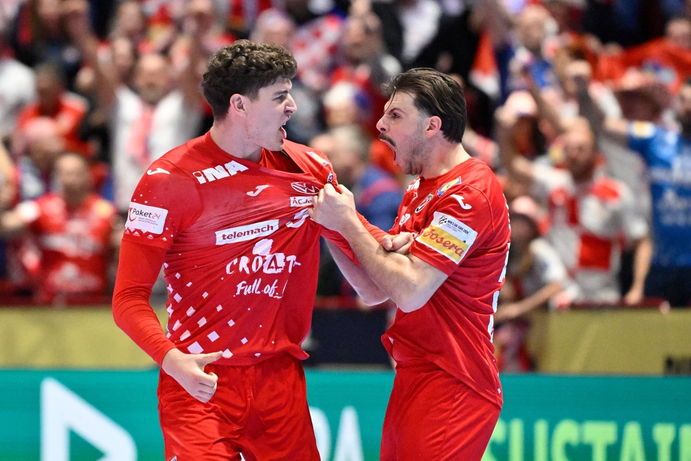 Dwóch mężczyzn w czerwonych strojach sportowych świętuje emocjonującą chwilę, prawdopodobnie po zdobyciu bramki podczas meczu piłki ręcznej. W tle widać widownię dopingującą sportowców.