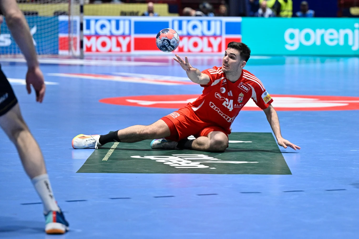 Zawodnik ubrany w czerwony strój leży na parkiecie podczas meczu piłki ręcznej i próbuje sięgnąć piłkę, w tle widoczne są reklamy oraz fragmenty boiska i inny zawodnik w czarnych spodenkach.