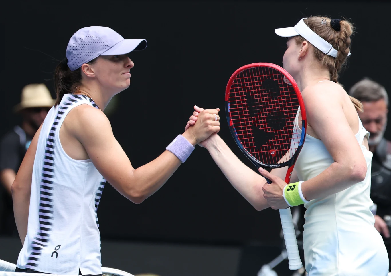 Dwie tenisistki podają sobie ręce przy siatce po meczu. Jedna z zawodniczek ubrana w białą koszulkę i czapkę z daszkiem, druga w jasnej sukience sportowej z białą opaską na głowie. W tle widoczny sędzia oraz kilka osób z obsługi turnieju.