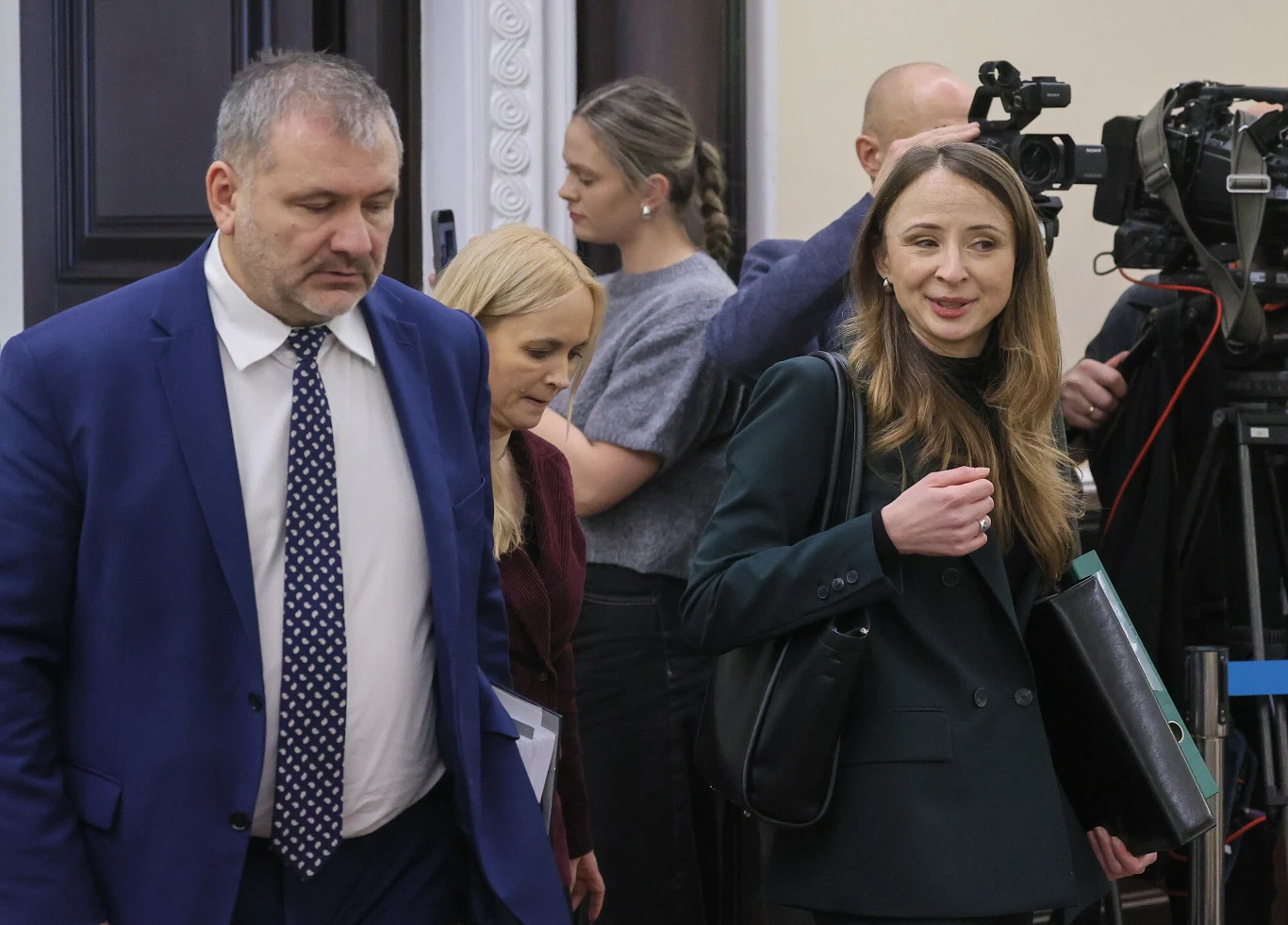 Wraca temat reformy PIP. Trwa wyścig z czasem, żeby nie stracić miliardów euro z KPO Grupa osób ubranych elegancko idzie korytarzem, z tyłu znajdują się kamery telewizyjne i dziennikarze przygotowujący się do relacjonowania wydarzenia. Atmosfera oficjalna, sugerująca uczestnictwo w spotkaniu lub konferencji.