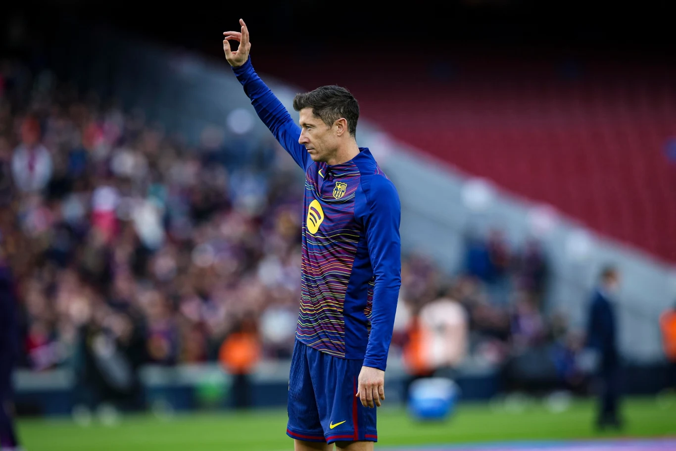 Piłkarz w stroju treningowym FC Barcelony unosi rękę ku górze, stojąc na boisku z rozmazaną publicznością w tle oraz stadionowym otoczeniem.