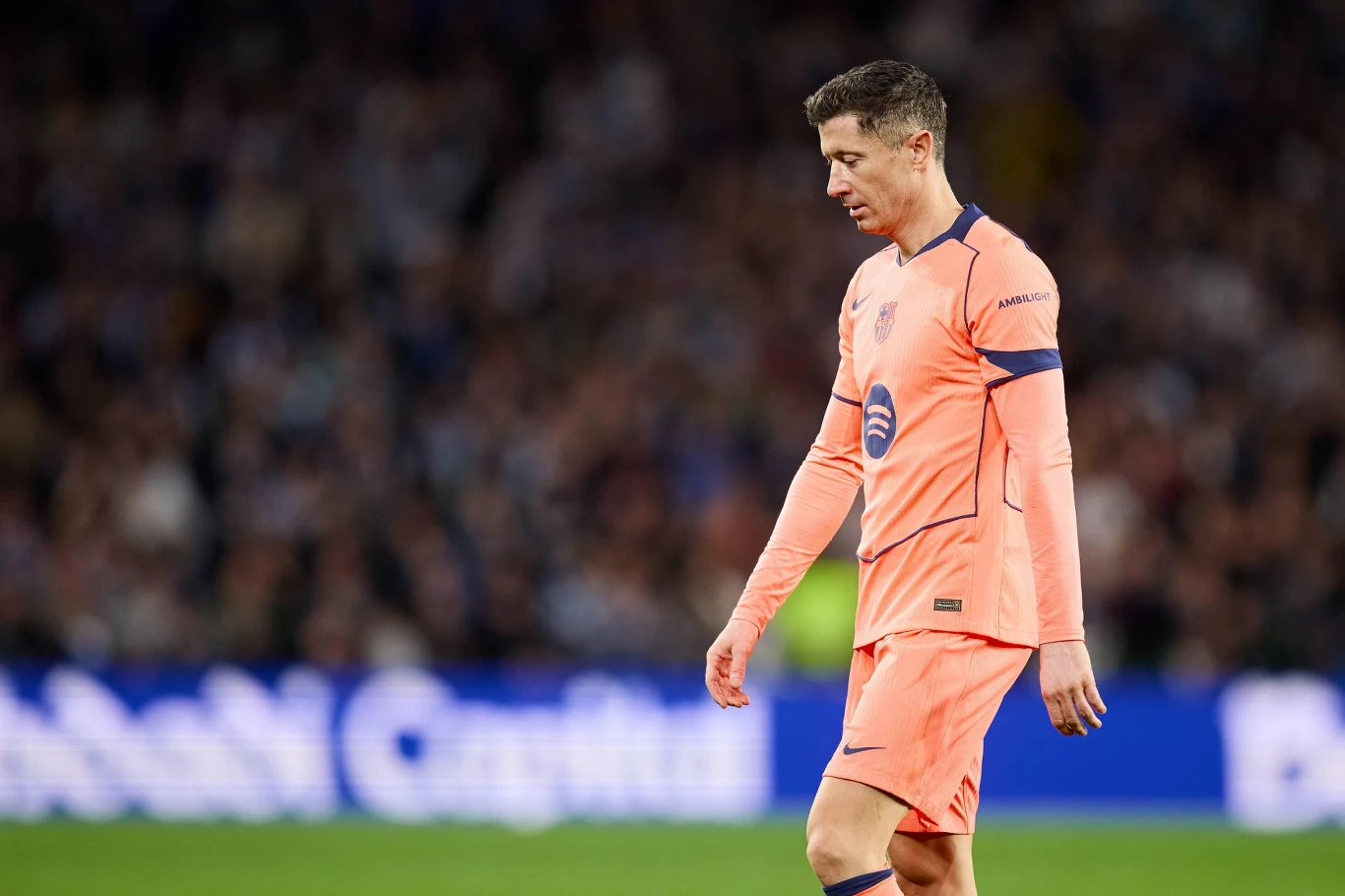 Piłkarz w pomarańczowym stroju drużyny FC Barcelona spaceruje po murawie podczas meczu piłki nożnej, w tle rozmyty tłum kibiców oraz reklamy stadionowe.