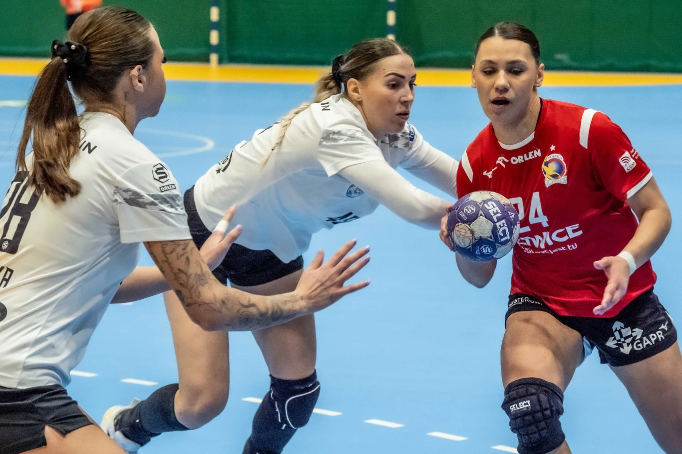 Trzy zawodniczki walczą o piłkę na parkiecie hali sportowej, dwie z nich w białych strojach próbują zatrzymać rywalkę w czerwonym stroju z piłką w rękach.