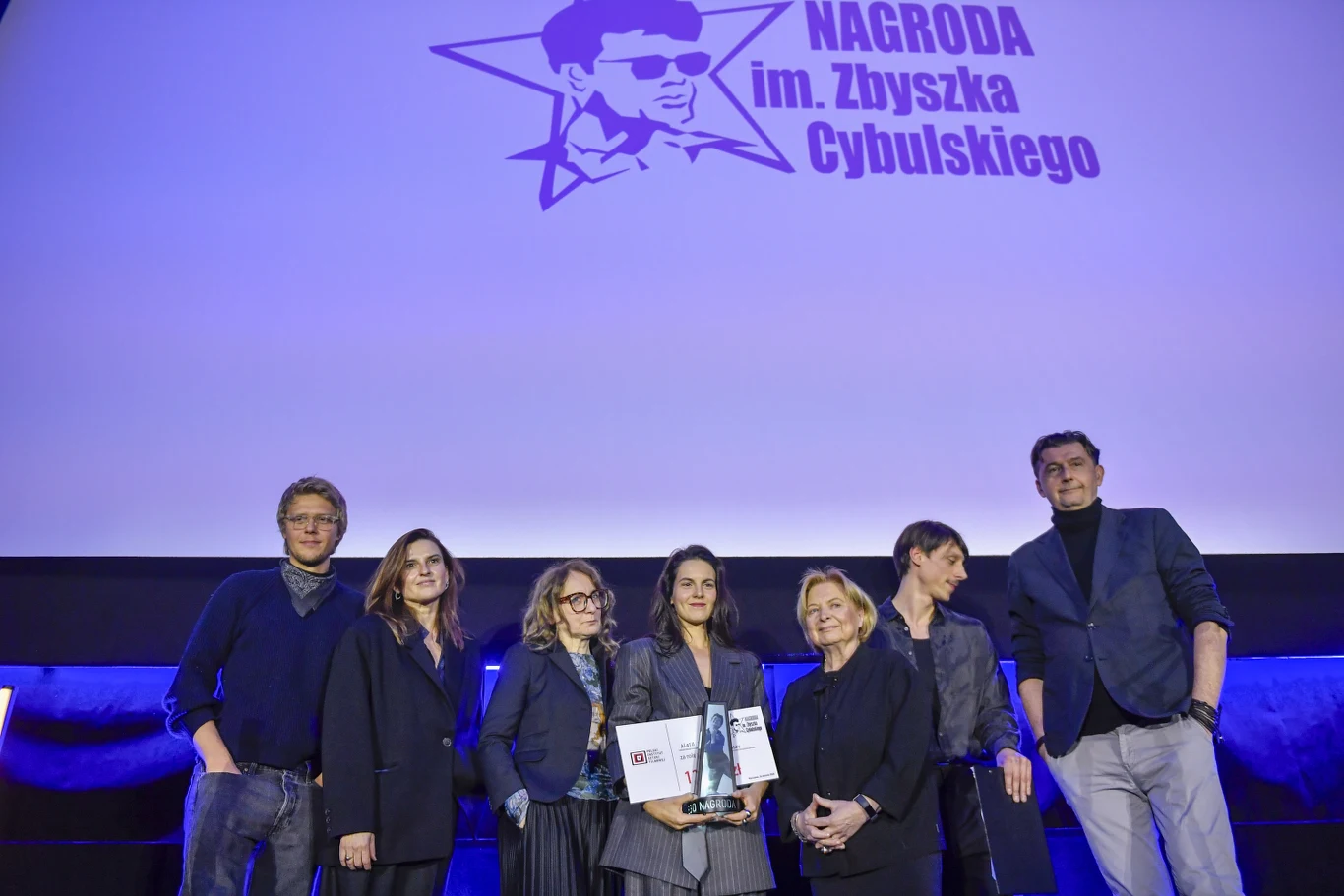 Grupa ludzi stojąca na scenie podczas ceremonii wręczenia nagrody imienia Zbyszka Cybulskiego, na tle dużej grafiki z nazwą nagrody. Jedna z osób trzyma statuetkę i dyplom.