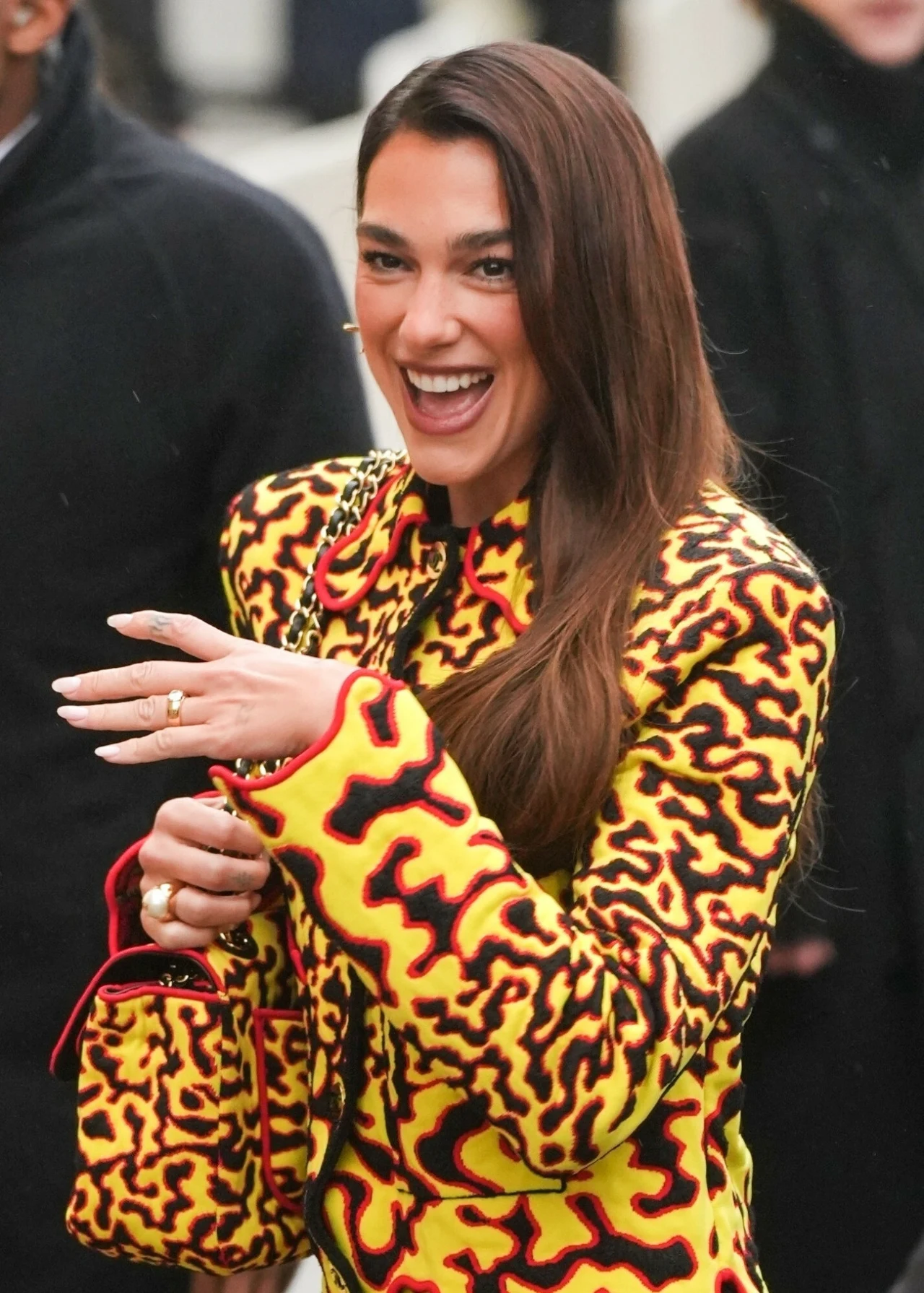 Kobieta z długimi włosami uśmiecha się szeroko, ubrana w żółtą kurtkę z abstrakcyjnym, czerwono-czarnym wzorem oraz trzyma dopasowaną kolorystycznie torebkę.