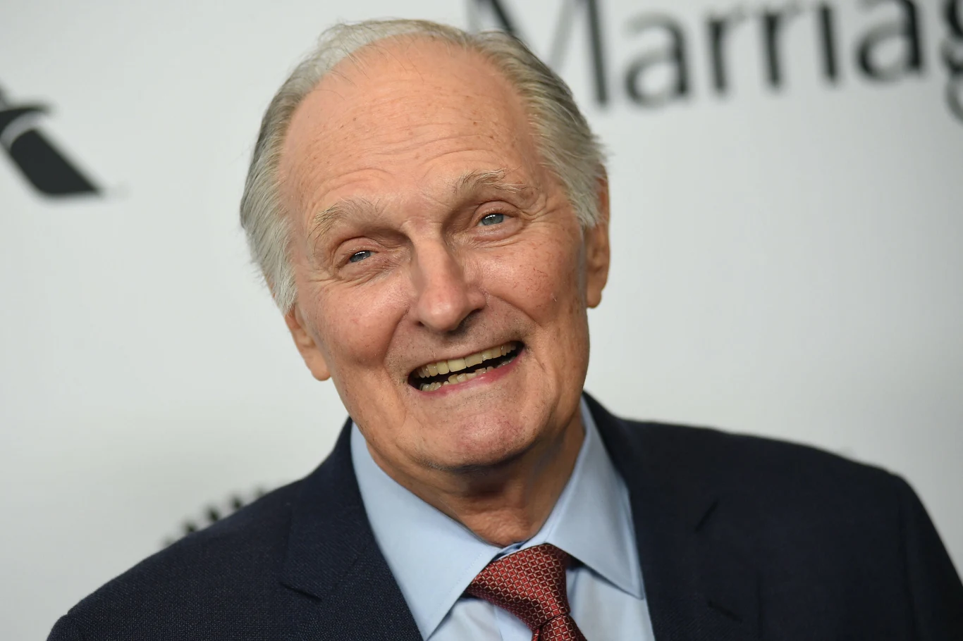 Elegancko ubrany starszy mężczyzna z szerokim uśmiechem na twarzy stoi na tle jasnej ścianki z częściowo widocznymi napisami, sugerując obecność na wydarzeniu branżowym lub kulturalnym.